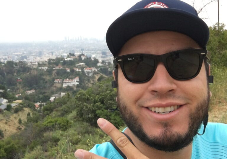 Luis wearing a turquoise shirt, hat and sunglasses, while giving the peace sign on a hill with houses and trees in the background